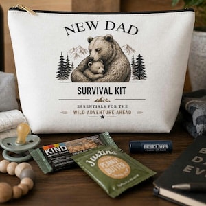 May include: A cream canvas wash bag with a black zip, printed with "NEW DAD SURVIVAL KIT" and a bear and cub design. Other items include a KIND bar, Justin's peanut butter sachet, Burt's Bees lip balm, a dummy, and a wooden teething ring.