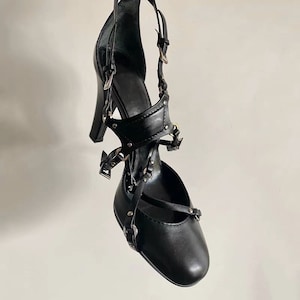 May include: Black leather heeled shoes with multiple straps and buckles. The straps are adorned with silver studs.