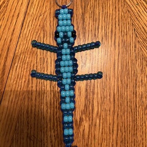 May include: A beaded lizard keychain in shades of blue and black. The lizard has a long body with four legs and a tail. The keychain is made of small, round beads strung together on a wire and attached to a silver keyring.