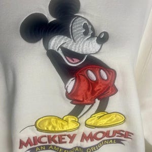 May include: White sweatshirt featuring an embroidered Mickey Mouse design. Mickey is depicted with black ears, a gray face, red shorts, and yellow shoes. The text "Mickey Mouse An American Original" is embroidered below the character.