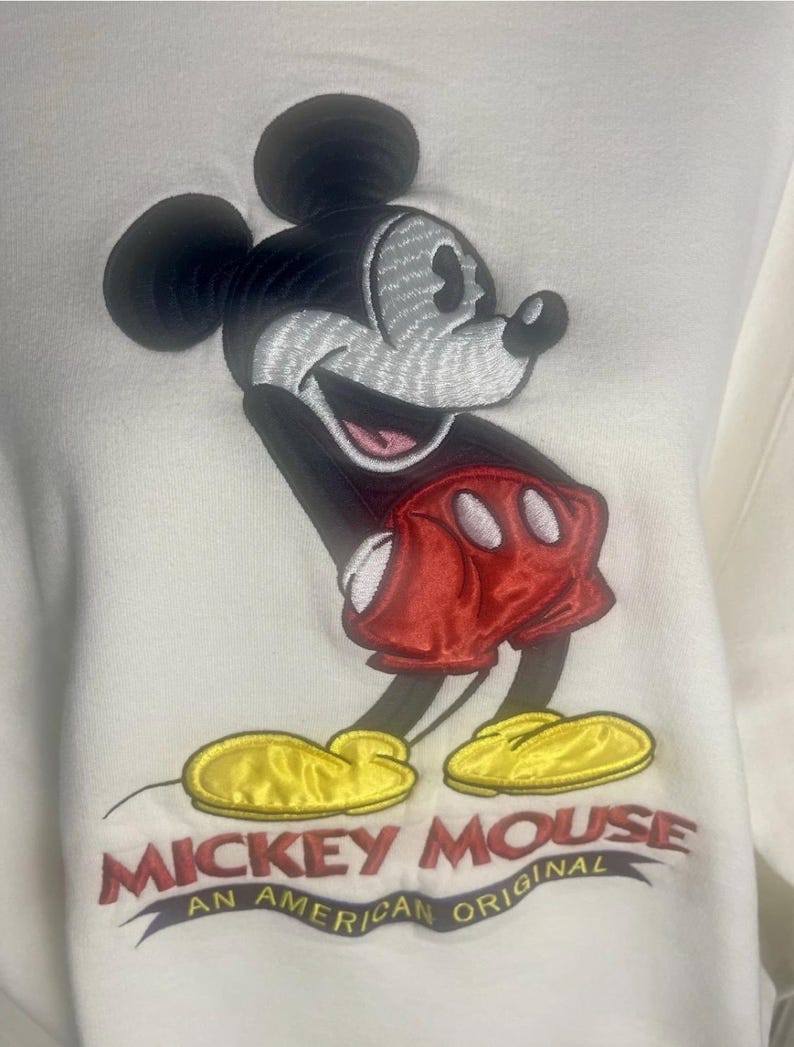 May include: White sweatshirt featuring an embroidered Mickey Mouse design. Mickey is depicted with black ears, a gray face, red shorts, and yellow shoes. The text "Mickey Mouse An American Original" is embroidered below the character.