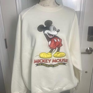 May include: Cream-colored sweatshirt featuring a large Mickey Mouse graphic. Mickey is depicted in black, red, and yellow, with the text "Mickey Mouse An American Original" below. The sweatshirt has long sleeves and a crew neck.