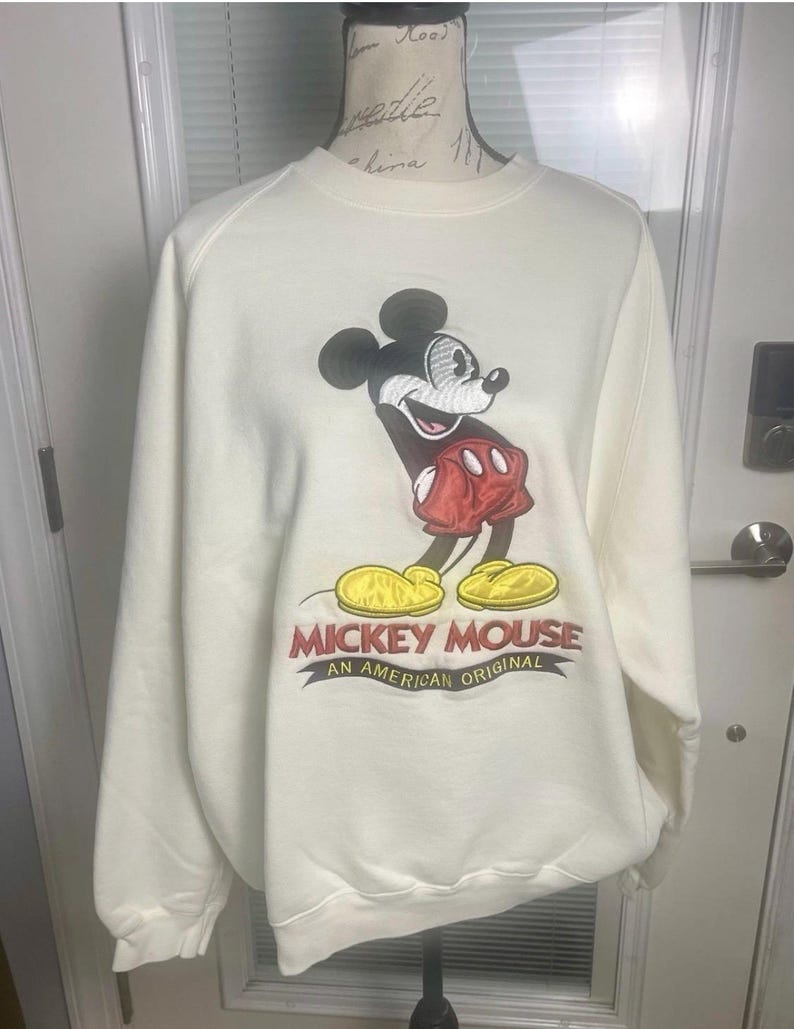 May include: Cream-colored sweatshirt featuring a large Mickey Mouse graphic. Mickey is depicted in black, red, and yellow, with the text "Mickey Mouse An American Original" below. The sweatshirt has long sleeves and a crew neck.