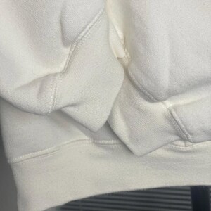 May include: Close-up of a cream-colored sweatshirt. The image highlights the texture and stitching details of the fabric, focusing on the cuffs and hem. The material appears soft and comfortable, suggesting casual wear.