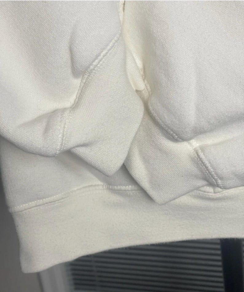 May include: Close-up of a cream-colored sweatshirt. The image highlights the texture and stitching details of the fabric, focusing on the cuffs and hem. The material appears soft and comfortable, suggesting casual wear.