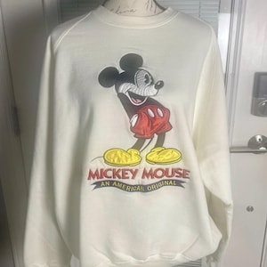 May include: Cream-colored sweatshirt featuring a large embroidered image of Mickey Mouse in his classic pose. Mickey is wearing red shorts and yellow shoes. The text "MICKEY MOUSE AN AMERICAN ORIGINAL" is below the image.