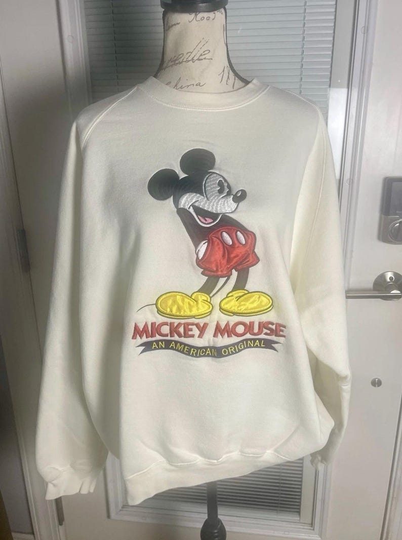 May include: Cream-colored sweatshirt featuring a large embroidered image of Mickey Mouse in his classic pose. Mickey is wearing red shorts and yellow shoes. The text "MICKEY MOUSE AN AMERICAN ORIGINAL" is below the image.