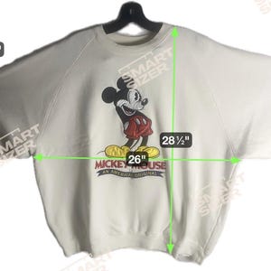 May include: Off-white sweatshirt featuring a Mickey Mouse graphic. The graphic shows Mickey Mouse in red shorts and yellow shoes. The sweatshirt is 26 inches wide and 28.5 inches long. The text "Mickey Mouse An American Original" is below the graphic.