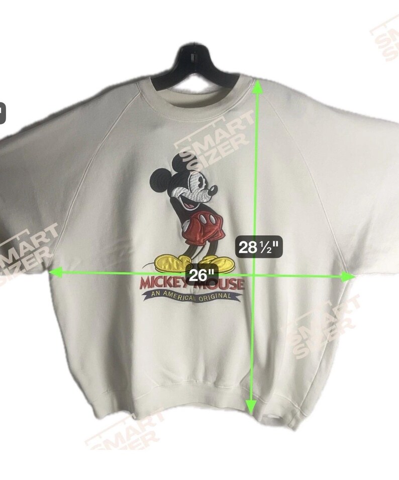 May include: Off-white sweatshirt featuring a Mickey Mouse graphic. The graphic shows Mickey Mouse in red shorts and yellow shoes. The sweatshirt is 26 inches wide and 28.5 inches long. The text "Mickey Mouse An American Original" is below the graphic.