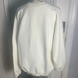 May include: Cream-colored sweatshirt with a crew neck and long sleeves. The back view of the garment is displayed on a mannequin. The sweatshirt has a simple, classic design, suitable for casual wear.