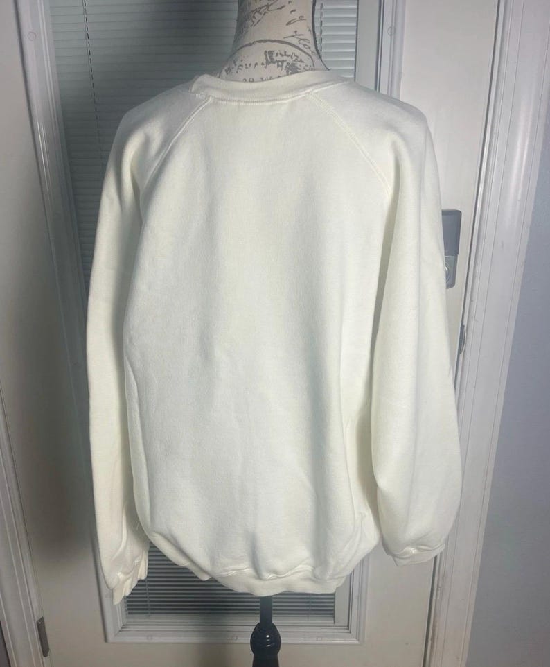 May include: Cream-colored sweatshirt with a crew neck and long sleeves. The back view of the garment is displayed on a mannequin. The sweatshirt has a simple, classic design, suitable for casual wear.