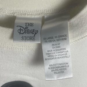 May include: Close-up of a cream-colored t-shirt with the Disney Store label. The label includes text such as "XX-LARGE" and "MADE IN SINGAPORE". The shirt is made of 80% cotton and 20% polyester.