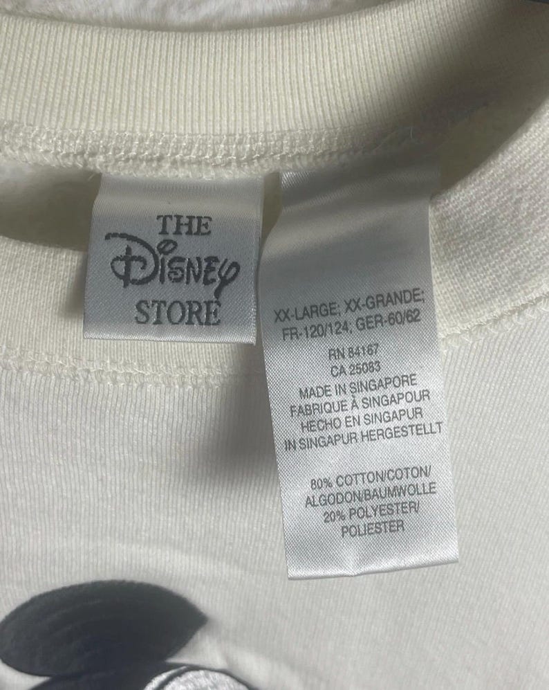 May include: Close-up of a cream-colored t-shirt with the Disney Store label. The label includes text such as "XX-LARGE" and "MADE IN SINGAPORE". The shirt is made of 80% cotton and 20% polyester.