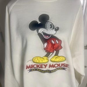 May include: Cream-colored sweatshirt featuring an embroidered image of Mickey Mouse. The design includes Mickey in his classic pose, wearing red shorts and yellow shoes. The text "MICKEY MOUSE AN AMERICAN ORIGINAL" is embroidered below the character.