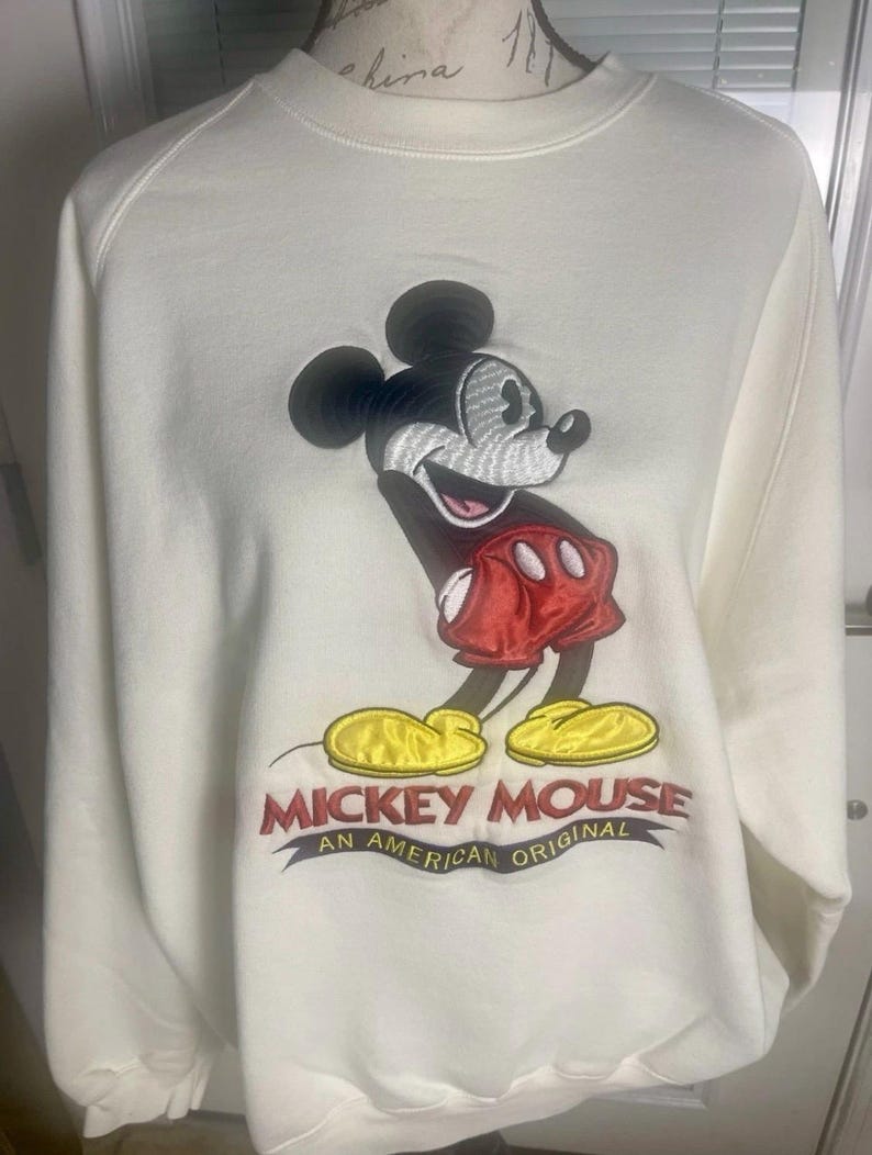 May include: Cream-colored sweatshirt featuring an embroidered image of Mickey Mouse. The design includes Mickey in his classic pose, wearing red shorts and yellow shoes. The text "MICKEY MOUSE AN AMERICAN ORIGINAL" is embroidered below the character.
