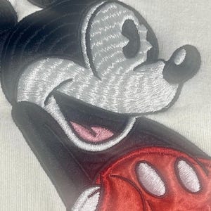 May include: Close-up of a white fabric with an embroidered Mickey Mouse design. The design features a black head and ears, a gray face, a red glove with white circles, and a pink mouth. The embroidery has a textured appearance.