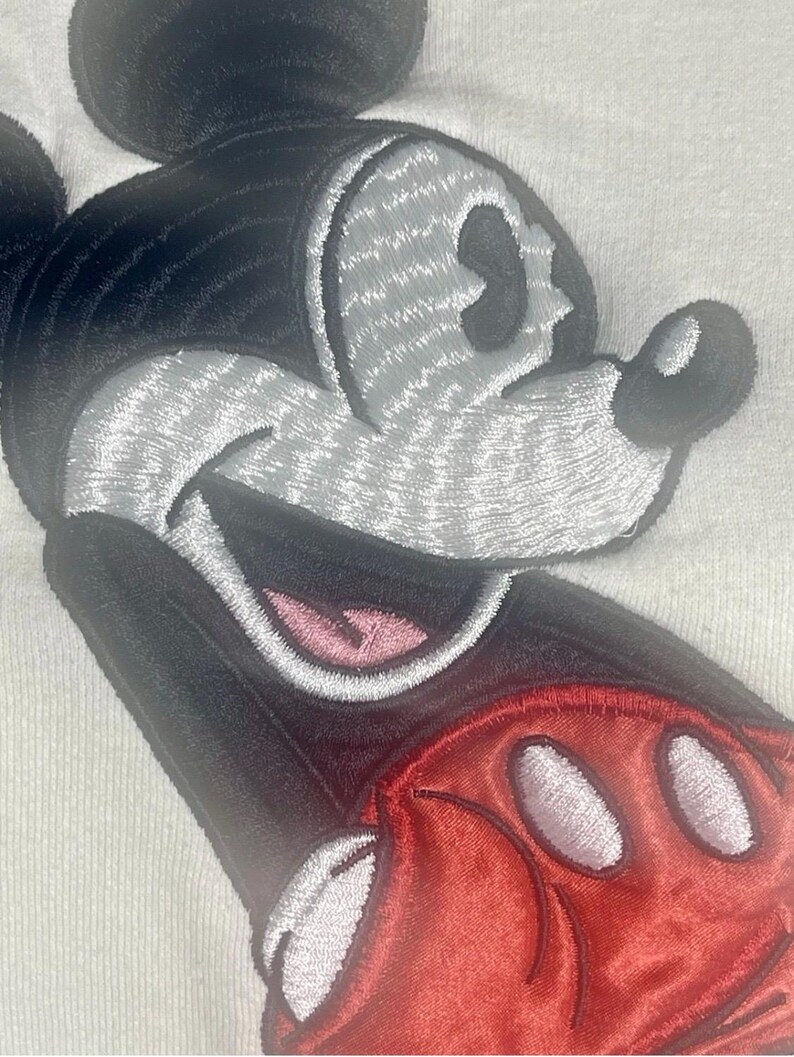 May include: Close-up of a white fabric with an embroidered Mickey Mouse design. The design features a black head and ears, a gray face, a red glove with white circles, and a pink mouth. The embroidery has a textured appearance.
