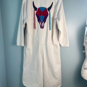 May include: Cream-colored long coat with a colorful bull skull design on the back. The bull skull features red, blue, and purple accents with matching fringe. The coat has long sleeves and a button-up front.