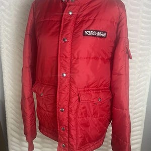Könnte beinhalten: Eine leuchtend rote, gesteppte Jacke mit Stehkragen und Druckknöpfen. Die Jacke hat zwei Vordertaschen und einen kleinen "YARD-MAN"-Aufnäher auf der Brust. Die Ärmel haben gerippte Bündchen.