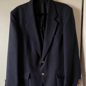May include: A dark navy blue blazer featuring a notched lapel, two gold-toned buttons at the center front closure, and three smaller matching gold-toned buttons on each sleeve cuff. The garment includes two flap pockets at the waist and a chest pocket.