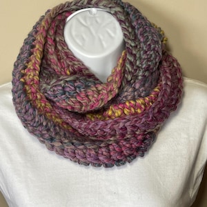 May include: A chunky knit infinity scarf in a blend of colors, including pink, purple, yellow, and gray. The scarf is displayed on a white mannequin head, showcasing its circular design and textured appearance. The yarn appears soft and warm.