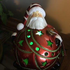 May include: A red and green Christmas ornament shaped like Santa Claus. The ornament has star-shaped cutouts that glow green. Santa's hat is striped red and white, and his beard is white. The ornament is on a dark surface.