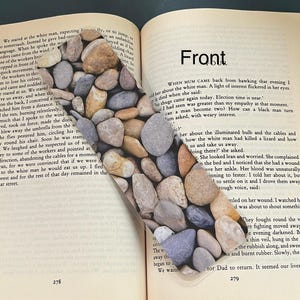May include: A rectangular bookmark featuring a high-resolution photographic print of smooth, multi-coloured river stones in shades of grey, tan, and brown. The bookmark is placed diagonally across the open pages of a book with printed text.