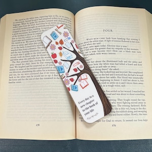 May include: A white bookmark with a brown tree illustration featuring colorful books, leaves, and flowers. The text reads, "Every day is a new chapter in the book of your life." The bookmark is placed diagonally across the open pages of a printed book.