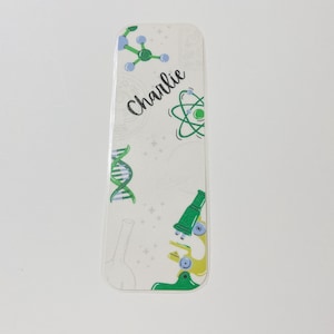 以下が含まれることがあります： 緑、青、白の科学をテーマにした白いしおり。しおりには、顕微鏡、DNA鎖、原子、分子のイラストが描かれています。しおりには、「チャーリー」という名前が黒い筆記体で書かれています。