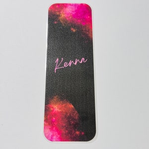 Puede incluir: Un marcapáginas de galaxia de color negro y rosa con el nombre "Kenna" escrito en rosa.