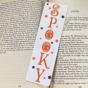 Könnte beinhalten: Ein weißes, rechteckiges Lesezeichen mit dem vertikal angeordneten, orangefarbenen Schriftzug SPOOKY. Das Design zeigt zwei orangefarbene Geisterillustrationen in kreisförmigen Rahmen sowie kleine orangefarbene Sterne, schwarze Sterne und winzige Spinnen.