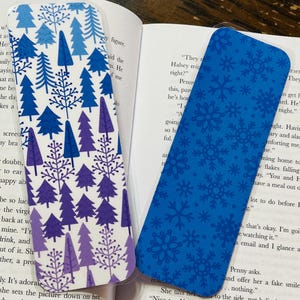 Puede incluir: Dos diseños de marcapáginas. Uno presenta un fondo azul con copos de nieve blancos. El otro presenta un fondo blanco con un patrón repetido de pinos estilizados de color morado y azul.