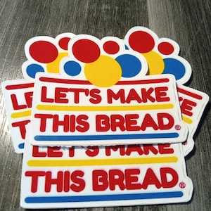 May include: A stack of white rectangular stickers with the text "LET'S MAKE THIS BREAD" in red, yellow, and blue. The stickers have a white border and are topped with colorful circles in red, yellow, and blue.