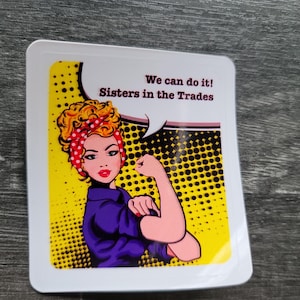 May include: A square sticker featuring a pop art illustration of a woman flexing her bicep, with the text "We can do it! Sisters in the Trades." The woman has red hair and a red polka dot headscarf, wearing a purple shirt.