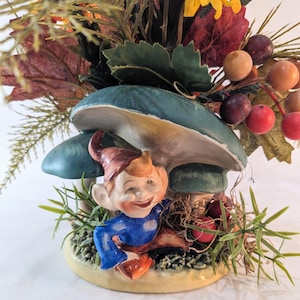 May include: A whimsical ceramic figurine featuring a smiling gnome in blue clothing and orange shoes, nestled under a cluster of teal mushrooms. The base is adorned with faux greenery and berries, with a backdrop of fall foliage.