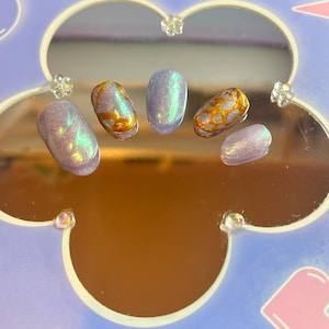 Może przedstawiać: Zestaw siedmiu iryzujących paznokci typu press-on w różnych kształtach. Paznokcie mają mieszankę odcieni lawendy, złota i zieleni. Niektóre paznokcie mają jednolity kolor, podczas gdy inne mają złoty, teksturowany wzór. Paznokcie są ułożone na lustrzanej powierzchni.