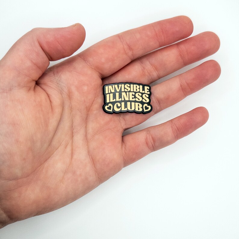 May include: Black enamel pin with the text "Invisible Illness Club" in gold lettering and two small gold hearts.