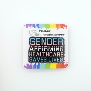 May include: A black enamel pin with the text "Gender Affirming Healthcare Saves Lives" in rainbow colors. The pin is on a white card with a rainbow border and the text "Ye Olde Queer Shoppe".