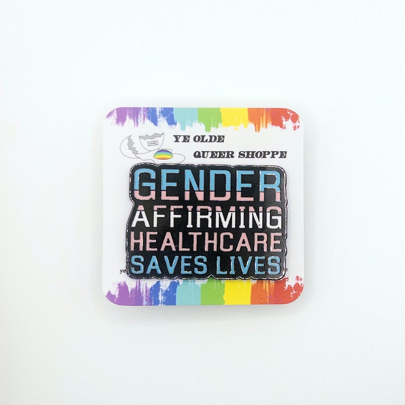 May include: A black enamel pin with the text "Gender Affirming Healthcare Saves Lives" in rainbow colors. The pin is on a white card with a rainbow border and the text "Ye Olde Queer Shoppe".
