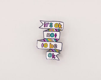 May include: A white enamel pin with a colorful banner that reads "it's ok not to be ok".
