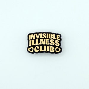 May include: Black enamel pin with the text "Invisible Illness Club" in gold lettering and two gold hearts.