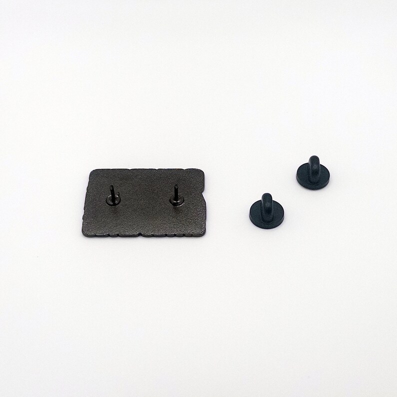 May include: The back of a black enamel pin with two metal pin backs. The pin features a design of a cartoon character with a speech bubble.