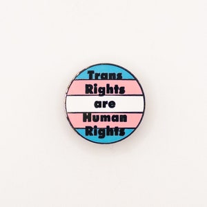 Peut inclure: Une épingle ronde avec les couleurs du drapeau de la fierté transgenre. L'épingle dit "Trans Rights are Human Rights" en texte noir.