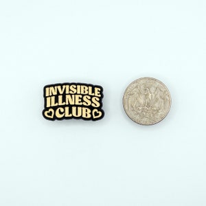 May include: Black and gold enamel pin with the text "Invisible Illness Club" in a retro font. The pin has two small heart shapes on either side of the word "Club".