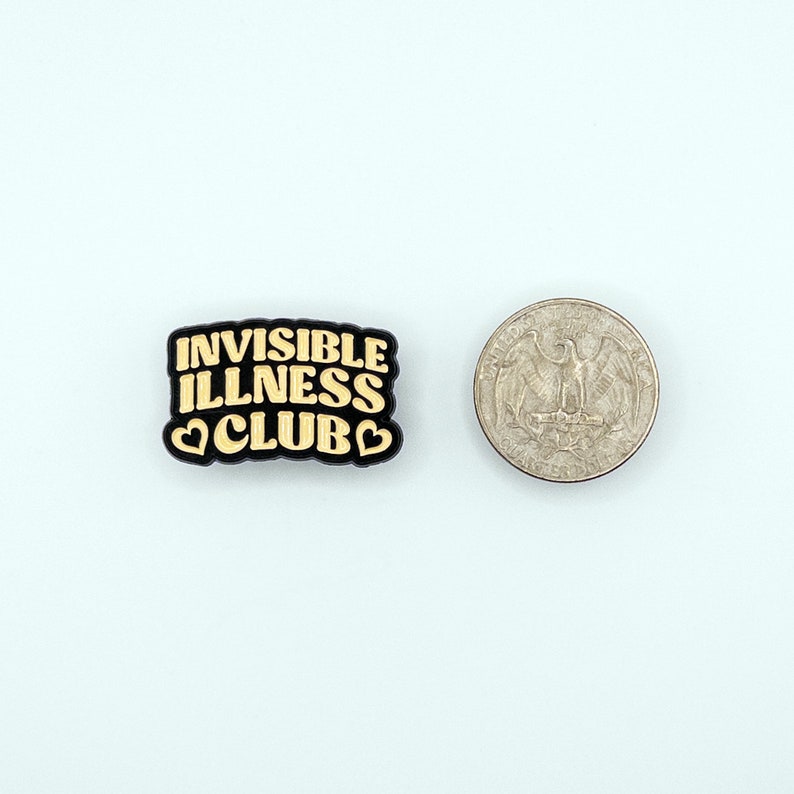 May include: Black and gold enamel pin with the text "Invisible Illness Club" in a retro font. The pin has two small heart shapes on either side of the word "Club".