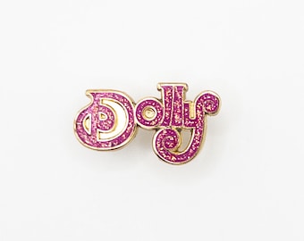 May include: A gold-colored pin with the word "Dolly" in pink glitter lettering.