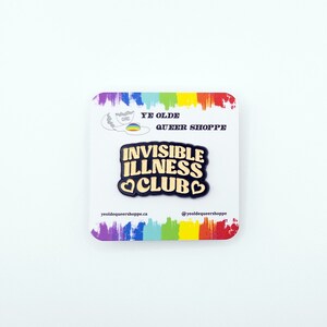 May include: Black enamel pin with gold lettering that reads "Invisible Illness Club" with a heart on each side of the word "Club". The pin is on a white card with a rainbow border and the text "Ye Olde Queer Shoppe" and the shop's website and social media handle.