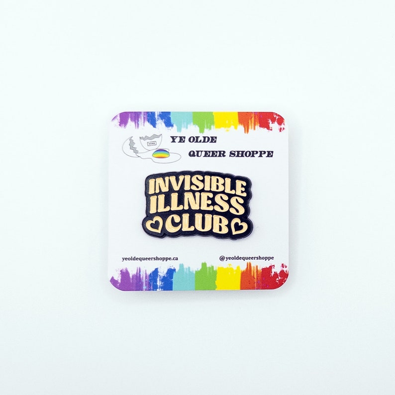 May include: Black enamel pin with gold lettering that reads "Invisible Illness Club" with a heart on each side of the word "Club". The pin is on a white card with a rainbow border and the text "Ye Olde Queer Shoppe" and the shop's website and social media handle.
