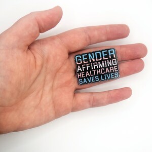 May include: A black enamel pin with the text "GENDER AFFIRMING HEALTHCARE SAVES LIVES" in white and blue lettering. The text is arranged in a stacked format with a pink and blue stripe between the words "GENDER" and "AFFIRMING".