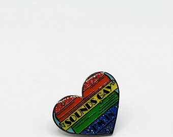 May include: A rainbow heart-shaped enamel pin with the text "Sounds Gay, I'm In" in black lettering.
